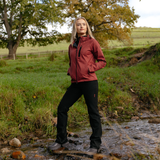 LeMieux AW26 Athena Waterproof Lightweight Jacket Varient Russet. A woman stands in a shallow outdoor stream wearing a muted red hooded jacket, black pants, and gray hiking boots, with grass, trees, and a fence in the background.