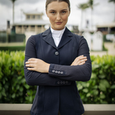 Woman in equestrian attire standing with arms crossed outdoors.