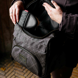 A person holds open a dark gray plaid grooming bag, showing its interior and a small black grooming brush inside. The bag has a front zippered pocket with a gold-toned 'LeMieux Heritage' logo patch.