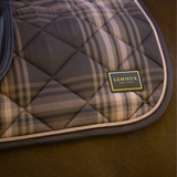 A close-up of a quilted horse saddle pad with a brown, beige, and gray plaid pattern, featuring a black leather brand patch that reads 'LEMIEUX HERITAGE'.