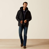 A man wearing a black hooded jacket over a navy shirt, blue jeans, and brown boots, standing against a plain beige studio background.