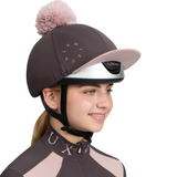 LeMieux AW26 Classique Pom Hat Silk Variant Colour Cocoa.
A woman wearing a cocoa colour equestrian helmet cover with a light pink pom-pom and matching visor. The cover features the brand name 'Le Mieux' in a circular design on the front, with a silver visor trim and black chin strap.