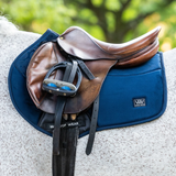 Woof Wear Elite Comfort Jump Saddle Pad Variant Navy. A brown leather equestrian saddle with a black stirrup, resting on a navy blue protective pad over a white horse's back, with a blurred green outdoor background.