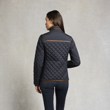 Holland Cooper Boughton Diamond Quilt Jacket Variant Colour Ink Navy.
A woman shown from the back wearing a dark navy quilted jacket with tan contrast stitching, paired with dark navy pants, against a textured light gray studio background.