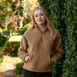 LeMieux AW26 Avery Teddy Quarter Zip Fleece Varient Caramel. A young woman with blonde hair standing outdoors in front of a leafy green hedge, wearing a tan fleece quarter-zip pullover with a small brown logo patch on the left chest.