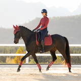 LeMieux AW26 Suede Dressage Square Varient Russet. A rider in equestrian apparel sits on a dark horse in an outdoor riding arena. The horse wears red protective leg wraps and a red saddle pad, with a matching red horse veil.