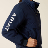 A close-up of a person wearing a navy blue jacket with large white vertical ARIAT branding on the left sleeve and a small circular logo on the left chest, against a plain light beige background.
