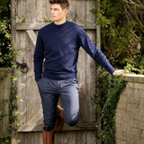 Man in navy sweater and the LeMieux Mens Elite Classic Pleated Breeches in Flint standing against a wooden gate with greenery.