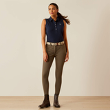 Woman wearing a navy sleeveless shirt, green riding pants, and brown boots on a beige background
