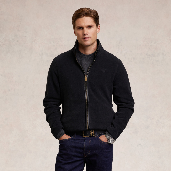 Holland Cooper Country Fleece Jacket (Men's) Variant Black. Man wearing a long sleeve black fleece with black leather trim detailing and a small HC logo on a beige background.