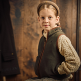 Holland Cooper Little Country Fleece Gilet Variant Khaki.A young girl wearing a brown knit beanie, a dark olive green zip-up vest over a cream cable-knit sweater, and a plaid skirt, posed against a rustic wooden background.