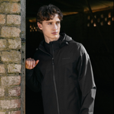 LeMieux AW26 Mens Waterproof Lightweight Jacket Varient Black. A man wearing a black hooded zip-up jacket stands in a doorway next to a brick wall, looking off-camera. The jacket has a subtle tonal logo on the left chest.
