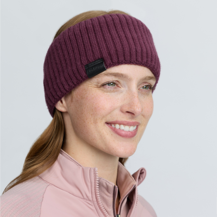 LeMieux AW26 Clara Rib Headband Variant Colouir Bramble.
A woman wearing a ribbed knit headband in a deep plum purple color, paired with a light pink zip-up jacket, against a solid light gray background.