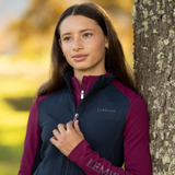 LeMieux AW26 Young Rider Bethany Fleece Zip Through Varient Blackberry. A woman outdoors leaning against a tree, wearing a LeMieux zip-front vest with dark navy body panels and deep burgundy sleeves and collar.