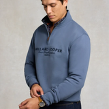 Holland Cooper Heritage Quarter Zip Sweat (Men's) Variant Denim Blue. Man wearing a blue 1/4 zip sweatshirt with Holland Cooper branding across the chest in navy agaisnt a grey background.