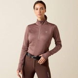 Ariat Ladies Team EQ 1/2 Zip Pullover Variant Twilight Mauve. A woman modeling a long-sleeve quarter-zip athletic top in a dusty mauve-brown color, paired with matching brown riding pants. The top features a small shield logo on the left chest and 'ABIA' printed subtly on the right sleeve.