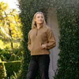LeMieux AW26 Avery Teddy Quarter Zip Fleece Varient Caramel. A woman wearing a tan fleece quarter-zip pullover sweater and black jeans stands outdoors in front of a white door framed by green ivy.