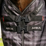 Close-up of a horse blanket with a gray and black plaid pattern, featuring black straps and plastic buckles, with the brand names 'LeMieux' and 'ARIKA' clearly visible.