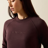 A close-up of a woman wearing a crewneck sweatshirt in a deep plum-brown color, featuring the brand name 'ARIAT' subtly embroidered on the chest.