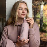 LeMieux AW26 Drinks Bottle Varient Wild Rose. A woman in a dusty pink hoodie holds a matching dusty pink LeMieux water bottle with a silver lid in an outdoor setting.