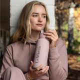 LeMieux AW26 Drinks Bottle Varient Wild Rose. A woman in a dusty pink hoodie holds a matching dusty pink LeMieux water bottle with a silver lid in an outdoor setting.