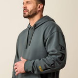 A man wearing a dark gray pullover hoodie with black embroidered lettering on the sleeve and a small yellow brand tag near the cuff, photographed against a plain light beige studio background.