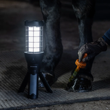 A black tripod-style portable lantern with a bright white light stands on a metal horse stall floor next to a horse leg, while a gloved hand applies liquid from an orange-handled brush to the hoof.