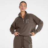 A woman modeling a dark brown quarter-zip sweatshirt and matching sweatpants set with a white circular logo on the chest and thigh, shot against a plain white studio background.