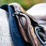 Woof Wear Elite Comfort Jump Saddle Pad Variant Navy. Close-up of a brown leather riding saddle with a dark navy blue padded under-saddle pad, resting on a horse's blanket.