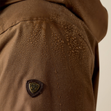 Close-up of a brown waterproof jacket with water droplets beaded on the fabric and a small shield-shaped logo patch on the shoulder.