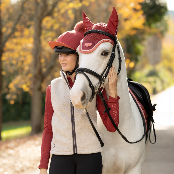 LeMieux AW26 Young Rider Trinity Teddy Gilet Varient Almond. A woman in equestrian apparel stands beside a white horse outdoors on a path with autumn trees in the background. The horse is wearing a red fly veil and black bridle, while the woman wears a cream fleece vest over a red long-sleeve top and black breeches.
