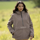 Reincoat Smock Lite Varient Dune. Woman wearing a Dune coloured jacket with a black zip and kangaroo style pocket with "REINCOAT" branding on a blurred green background.