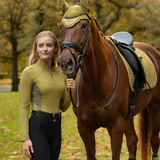 LeMieux AW26 Classique Base Layer Variant Colour Gooseberry.
A woman in equestrian apparel stands beside a brown horse outdoors. She wears a fitted gooseberry green long-sleeve riding shirt and black riding pants, while the horse is outfitted with a matching olive-green and black bridle and saddle pad.