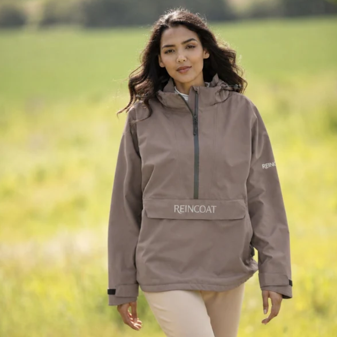Reincoat Smock Lite Varient Dune. Woman wearing a Dune coloured jacket with a black zip and kangaroo style pocket with "REINCOAT" branding on a blurred green background.