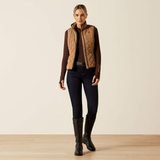 Woman wearing a brown quilted vest, dark jeans, and black boots on a beige background