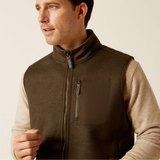 Man wearing a brown gilet over a beige shirt against a plain background