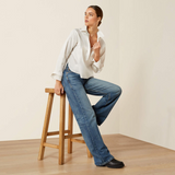 Woman in white shirt and blue jeans sitting on a wooden stool against a plain background