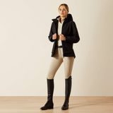 Woman wearing a black equestrian jacket, beige pants, and black boots on a plain background