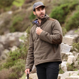 Man wearing a brown fleece jacket and cap standing in a natural setting with rocks and greenery.