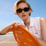 Female lounges on the beach in an orange chair wearing the Barbour Ladies Bowford Sleeveless Polo Top in White and a pair of dark, oval sunglasses