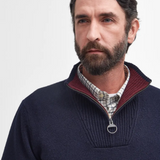 Man wearing a navy jumper with a red inner collar and plaid shirt underneath on a light grey background