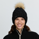LeMieux AW26 Bernie Sequin Beanie Variant Colour Black.
A smiling woman wearing a black cable-knit beanie with a fluffy tan pom-pom and subtle silver glitter, paired with a black fleece zip-up jacket against a light gray background.