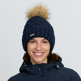 LeMieux AW26 Bernie Sequin Beanie Variant Colour Navy.
A smiling woman wearing a navy blue cable-knit beanie with a large tan faux-fur pom-pom and a small visible LeMieux logo, paired with a navy hooded jacket against a light gray studio background.
