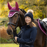 LeMieux AW26 Clara Knit Beanie Navy One Size Variant Colour Navy.
A woman in a navy equestrian jacket and navy beanie with a pom-pom stands beside a brown horse wearing a magenta fly veil and bridle in an outdoor setting with autumn trees.