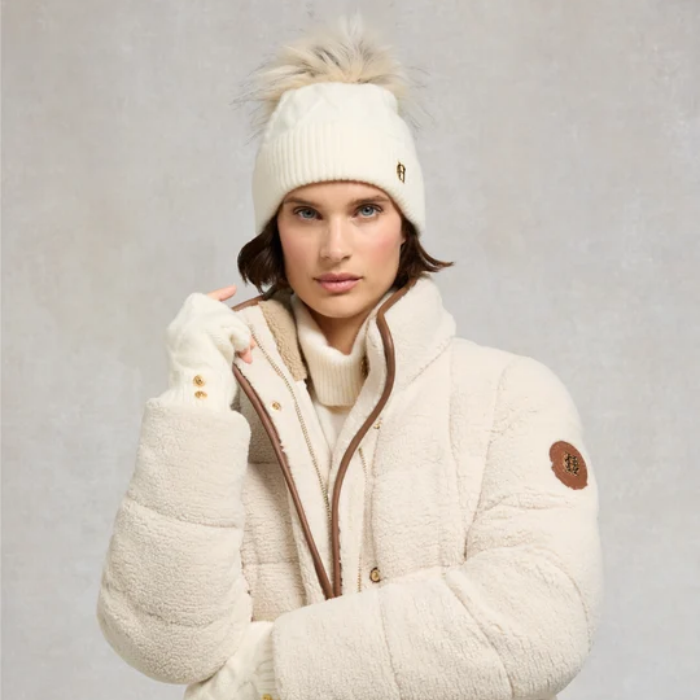 Lady wearing a cream bobble hat with a cream jacket against a grey background.