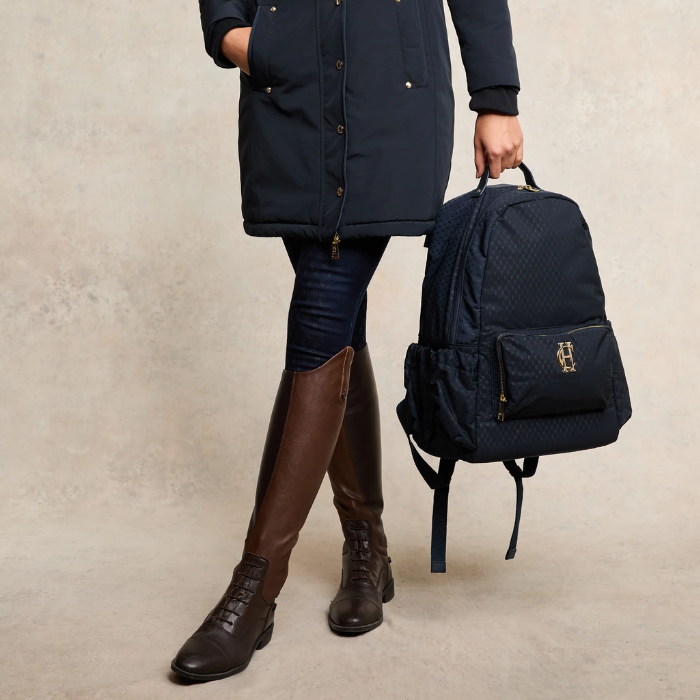 Holland Cooper Burghley Backpack Variant Ink Navy Monogram. A model wearing a dark navy coat, dark jeans, and brown knee-high boots holds a dark navy quilted backpack with gold hardware against a textured beige studio background.