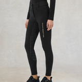 Holland Cooper Chiltern Contour Breeches Variant Black. Close up of someone wearing a pair of black breeches with Holland Cooperdown the side against a grey background.