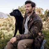 Man in a camouflage jacket sits with a dog on a rural hillside