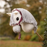 LeMieux AW26 Hobby Horse Vogue Headcollar & Leadrope Variant Colour Blackberry.
A plush toy horse head mounted on a stick, with a white body, gray faux-fur mane, and blackberry halter, photographed outdoors against a blurred garden background.