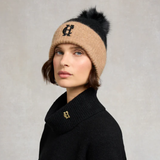 Woman wearing a beige knit beanie with black fur pom-pom and black coat with brand logo on a neutral background